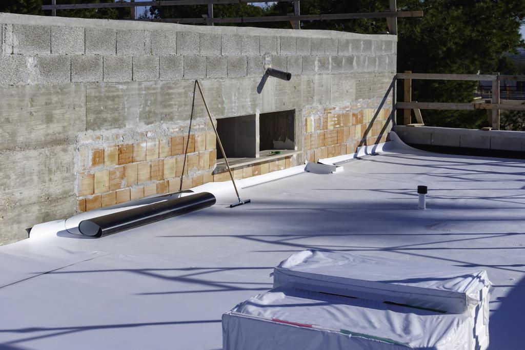 A flat rooftop under construction showcases a white TPO membrane partially installed. Rolls of material and a pipe jut from the roof. Concrete and brick walls encircle the site, with trees visible in the background.