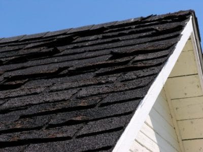 Close-up of a house roof with dark, weathered shingles showing signs of curling and damage. The sky is clear and blue, and part of the white siding is visible beneath the roofline, hinting at the need for expert care from Vancouver roofing services.