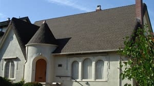 A quaint house with a steep, dark shingled roof crafted by expert roofing contractors in Richmond features a round turret with a conical top. The cream-colored exterior has arched windows and a wooden front door. A small tree is partially visible in the foreground.