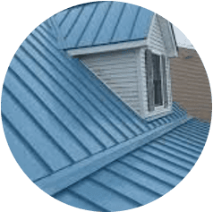 A blue metal roof with a gabled design and an upper-level window shows the expertise of commercial roofing services. The siding is light gray, contrasting beautifully with the roof. The angle captures the cladding and dormer window perfectly.
