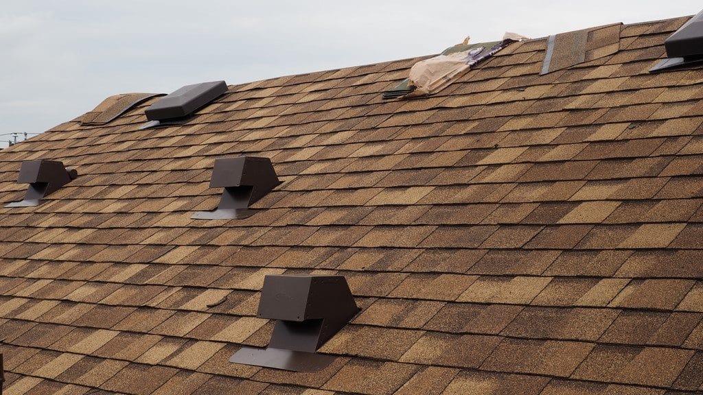 A sloped roof with brown asphalt shingles features multiple dark brown vents. One section, likely part of commercial jobs, has a package of shingles placed on it, indicating ongoing or recent roofing work. The sky remains overcast.