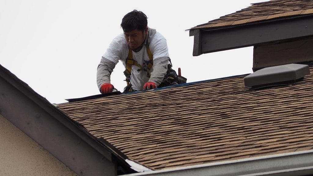 A worker in a white shirt and safety harness repairs brown roof shingles, using tools from his belt. Kneeling on the roof, focused on his task under an overcast sky, he demonstrates the skill and dedication found in commercial careers.