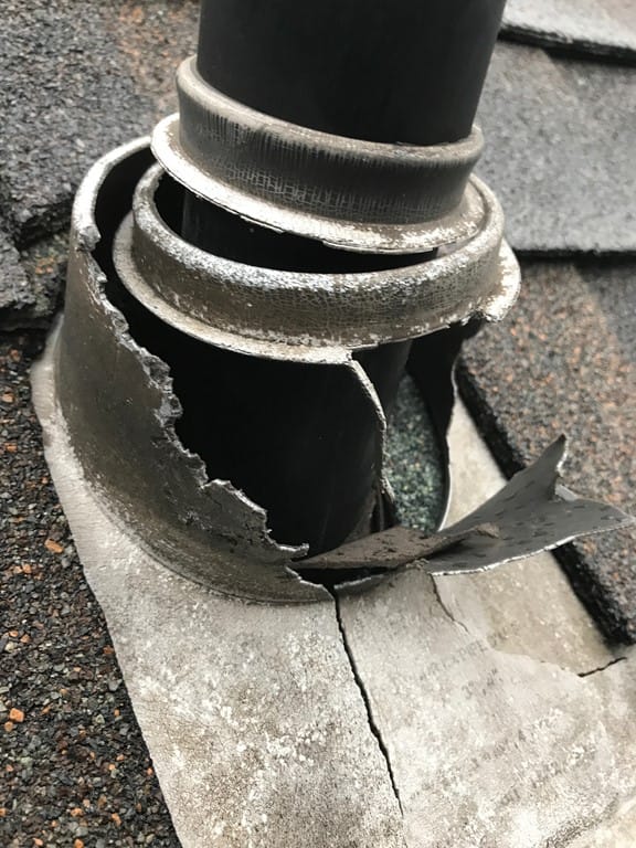 Damaged metal chimney flashing on a rooftop. The corroded and split best flashings reveal a black pipe underneath. Asphalt shingles surround the area.