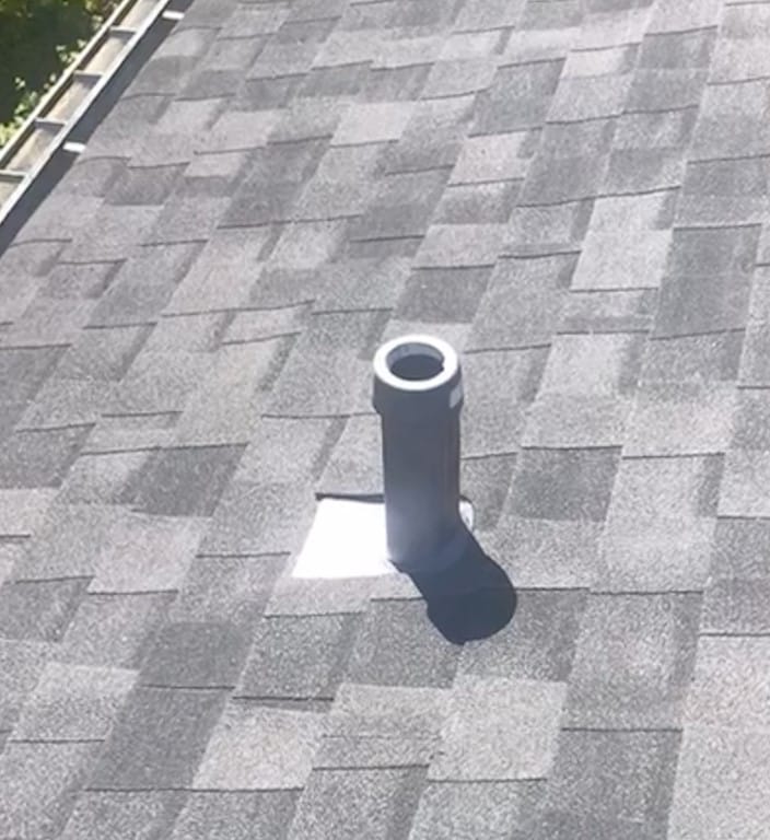A rooftop with grey asphalt shingles features a small, cylindrical chimney pipe with the best roof flashings surrounding it. Sunlight casts a shadow of the pipe on the shingles, while trees are partially visible in the background.