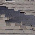 A roof with missing and damaged asphalt shingles screams for attention, as the black underlayment peeks through ominously. Shingles appear discolored and haphazardly arranged, signaling wear. A small vent sits near the top. Its time to replace your asphalt shingle roof before further damage occurs.