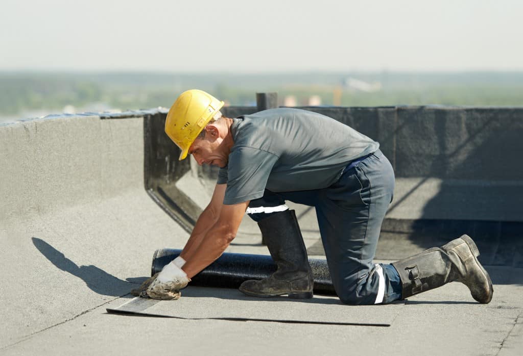 A construction worker in a yellow hard hat and gray clothing is kneeling on a flat rooftop, focused on roof maintenance. Using protective gloves, he carefully installs roofing material. The background is blurred, with hints of greenery on the distant horizon.