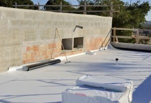 A construction site with a flat white roof in progress showcases building materials scattered about, including large rolls and square packages. A partially built brick and concrete wall stands tall, with trees and a wooden railing adding a natural backdrop.
