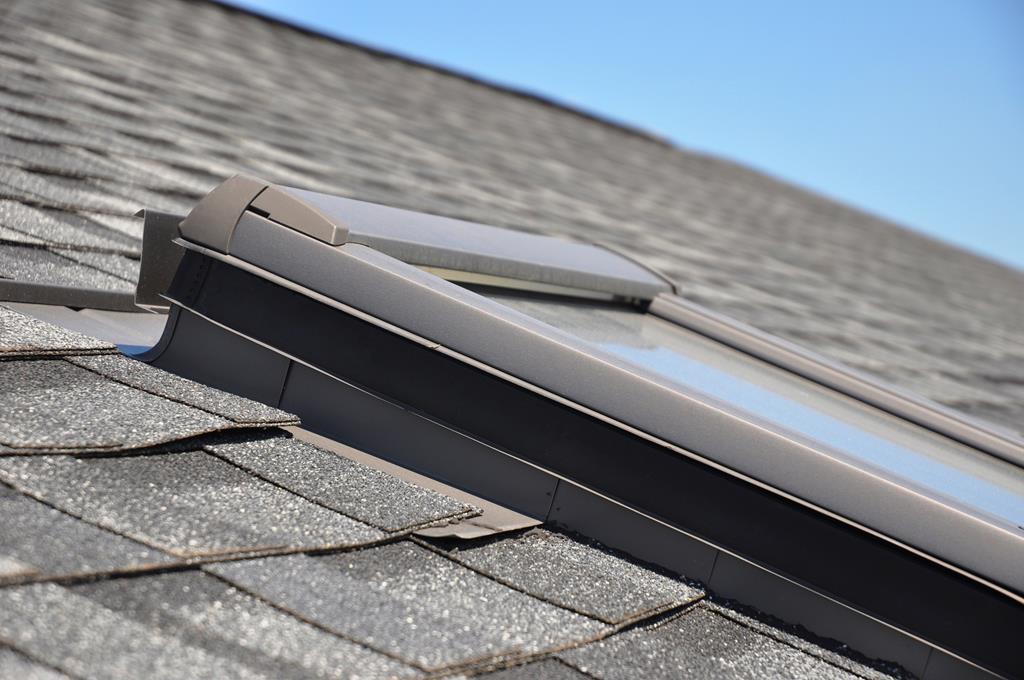 A close-up of a roof with gray asphalt shingles showcases the sleek skylight, its metal frame gleaming against the clear blue sky in the background.