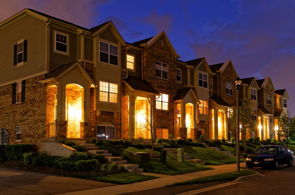 A row of Strata Properties townhouses features illuminated porches at dusk, their brick exteriors and peaked roofs silhouetted against a sky that mixes dark blue and orange. A few cars are parked along the street, completing this serene sunset scene.