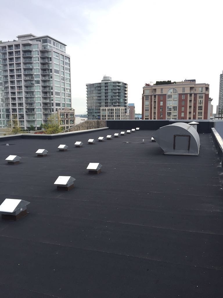 Rooftop view with evenly spaced vents on a black surface suggests an old commercial roof in need of repair. In the background, modern high-rise buildings rise against a cloudy sky, one featuring a large, curved structure.
