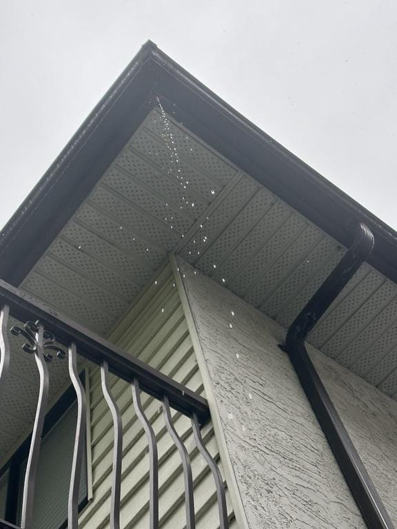 A view from below of a buildings corner features water droplets cascading from the roof, highlighting the importance of preventative roof maintenance. The structure boasts decorative wrought iron railing against a light-colored wall, set against an overcast sky that hints at rain.