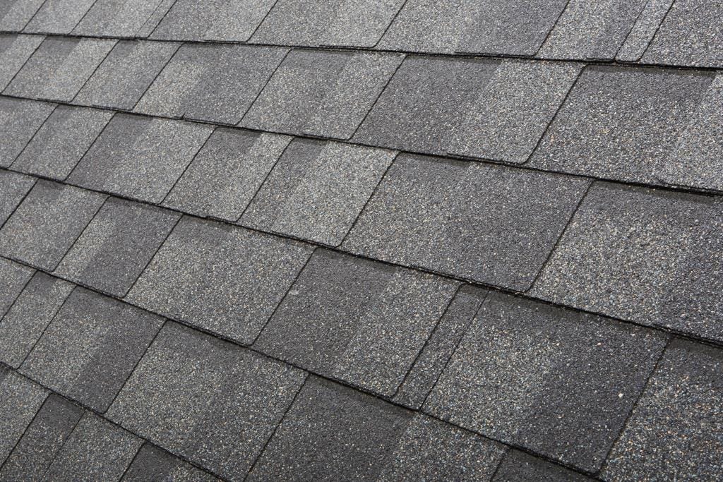 Close-up of a roof covered with overlapping dark gray asphalt shingles, demonstrating the importance of shingle quality. The neatly arranged shingles display a textured surface with granules, ensuring a durable and weather-resistant finish.