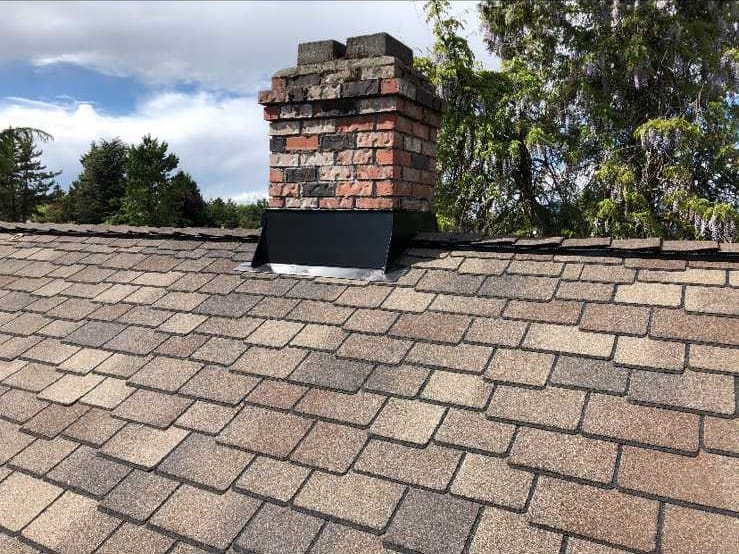 A sloped roof with brown shingles of high quality features a brick chimney with metal flashing. Trees and a partly cloudy sky are visible in the background, demonstrating an understanding of durable shingle grades.