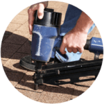 A person using a blue nail gun to install brown shingles on a roof as part of residential roofing services. The hands firmly grip the tool, and the textured surface is evident. Partially visible knees suggest kneeling during the task.