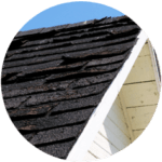 Close-up of a triangular house facade with worn, dark shingles showcasing signs of damage, hinting at the need for residential roofing services. The white wooden trim contrasts with the dark roof under a clear blue sky.