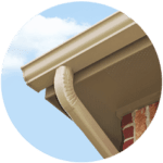Close-up of a beige gutter system attached to the corner of a building with red brickwork, showcasing quality residential roofing services. The sky is clear with a few clouds in the background. The gutter features a downspout leading from the roofline.