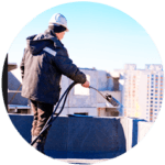 A construction worker in a hard hat and warm jacket expertly applies material to a rooftop using a long-handled tool, showcasing the precision of commercial roofing services against a blurred city skyline under the clear blue sky.