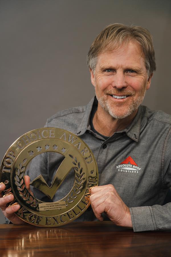 A man, representing Vancouver Roofing, smiles while seated at a table, proudly holding a gold Top Choice Award plaque. He wears a grey shirt adorned with the companys red and white logo against a plain backdrop.