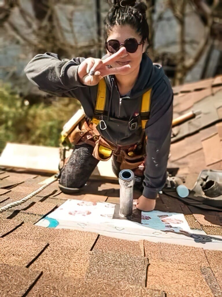 A woman in a grey sweatshirt, overalls, and sunglasses kneels on a rooftop, flashing a peace sign with one hand. Tools are scattered around her as she works on an area with a pipe and roofing material.