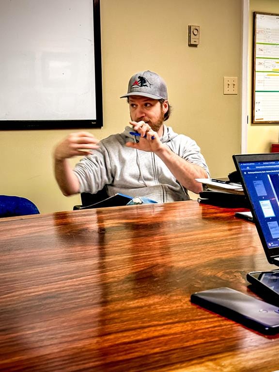 A person sits at a wooden table, wearing a gray hoodie and cap from Absolute Roof Solutions, gesturing with one hand while holding a pen in the other. A laptop is open on the table, emphasizing their Safety First approach, and a whiteboard is visible in the background.