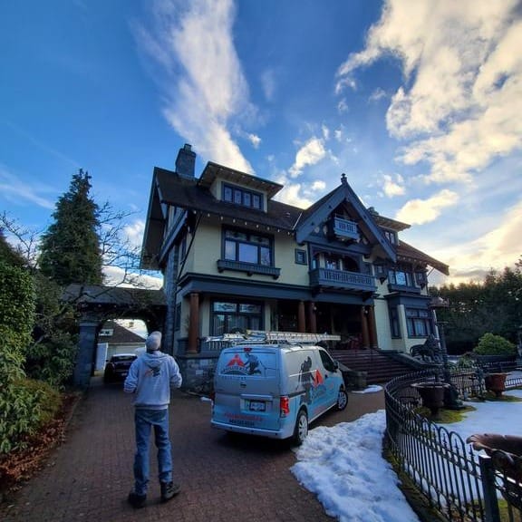 A roofer stands in the driveway of a large, elegant house with a steep roof, perfect for roof inspections. Attic windows peek out while a white van with a colorful logo rests nearby. Snow partially covers the ground as the sky shows blue hues with scattered clouds, all capturing the essence of protecting your investment.