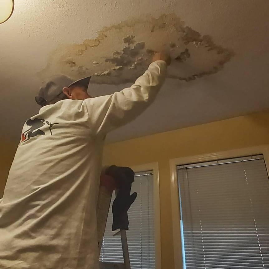 A person wearing a cap and long-sleeve shirt stands on a ladder, expertly repairing a water-damaged ceiling in a room with yellow walls and closed blinds. Their precision reflects why they receive 5-star reviews for their roof solutions.