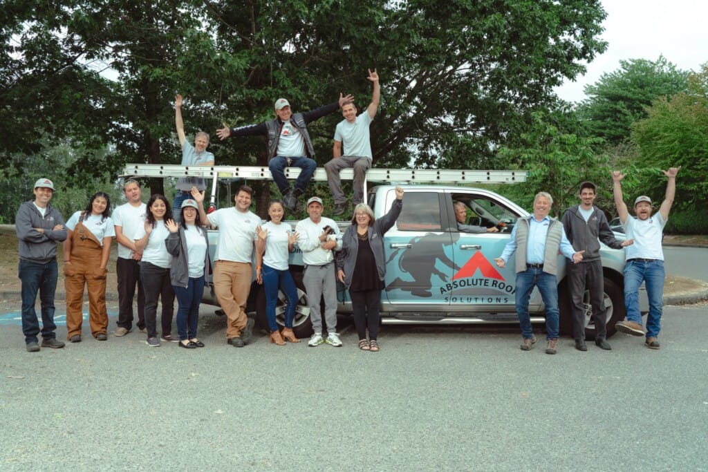 A group of satisfied clients poses energetically around a truck labeled Absolute Roof Solutions. Some are on the vehicle or truck bed. They are outdoors, with trees in the background, all wearing casual attire, celebrating their 5-star reviews.