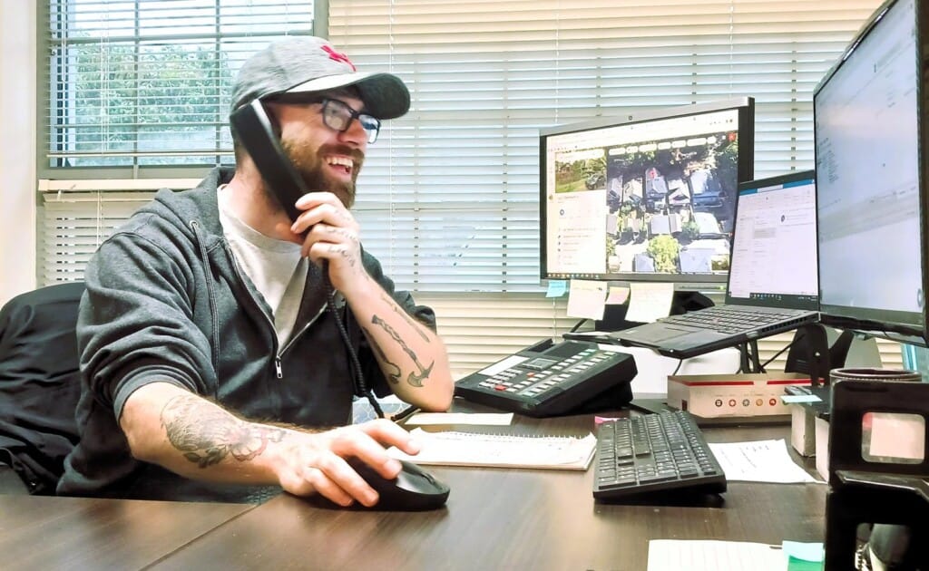 A bearded man with glasses and a cap is smiling while talking on a phone in an office, ensuring a great client experience. Hes using a computer mouse near the keyboard. Two monitors, a calculator, and sticky notes clutter the desk as light filters through partially open blinds.
