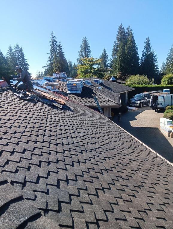 A person wearing safety gear is working on a sloped rooftop covered with dark asphalt shingles, ensuring no installation mistakes are made. Various materials and tools are scattered around. Several vans and parked cars fill the driveway below, while tall trees line the background under a clear sky.