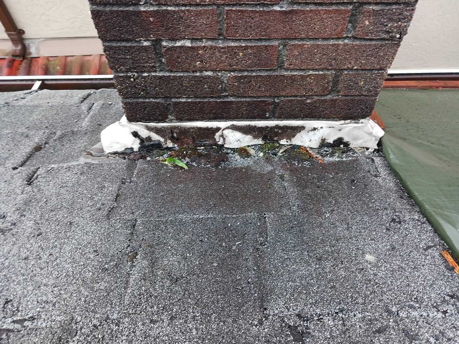 Close-up of a brick chimney base on a shingled roof, with white sealant around the edges and debris like leaves at the base—common roofing deficiencies that Vancouver home inspectors look for during a home inspection, highlighting possible water damage or leaks.