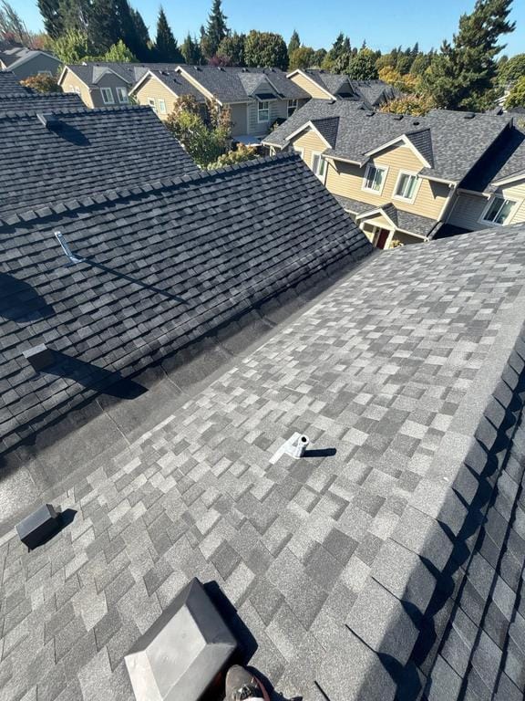 View from a rooftop showing gray asphalt shingles, vent pipes, and nearby houses with similar roofing—possibly part of a housing cooperative—surrounded by trees under a clear blue sky, ideal for an Absolute Roof Solutions roof replacement.