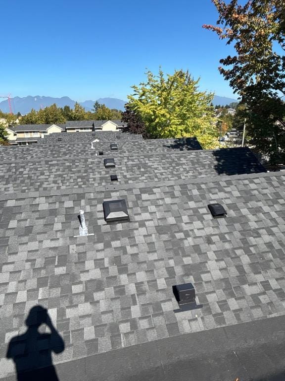 Shingled rooftop with multiple vents and pipes, surrounded by trees and neighboring houses, with mountains visible under a clear blue sky in the background. The scene suggests the quality work of Absolute Roof Solutions on a potential roof replacement project.