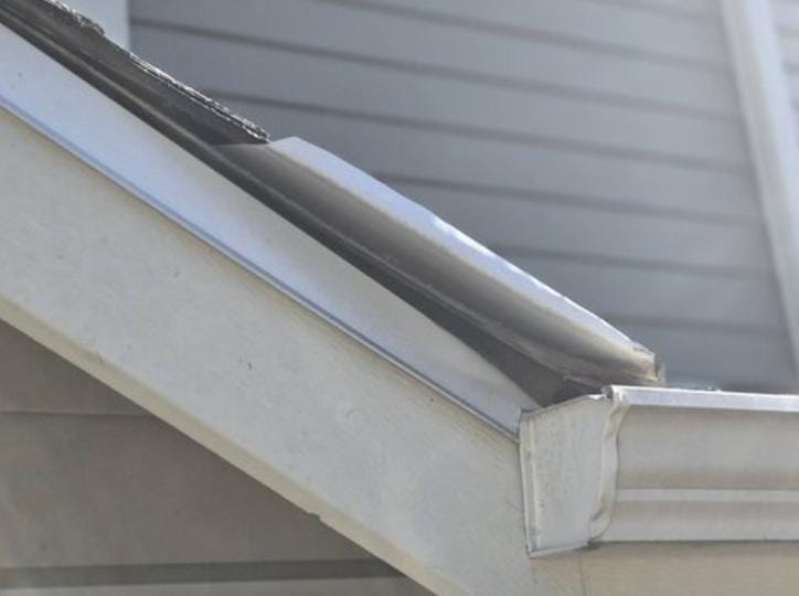 Close-up of a damaged house roof with shingles bent upward and detached near the edge, above a white gutter—an issue that may require roof replacement. Gray siding is visible in the background. Photo courtesy of Absolute Roof Solutions.